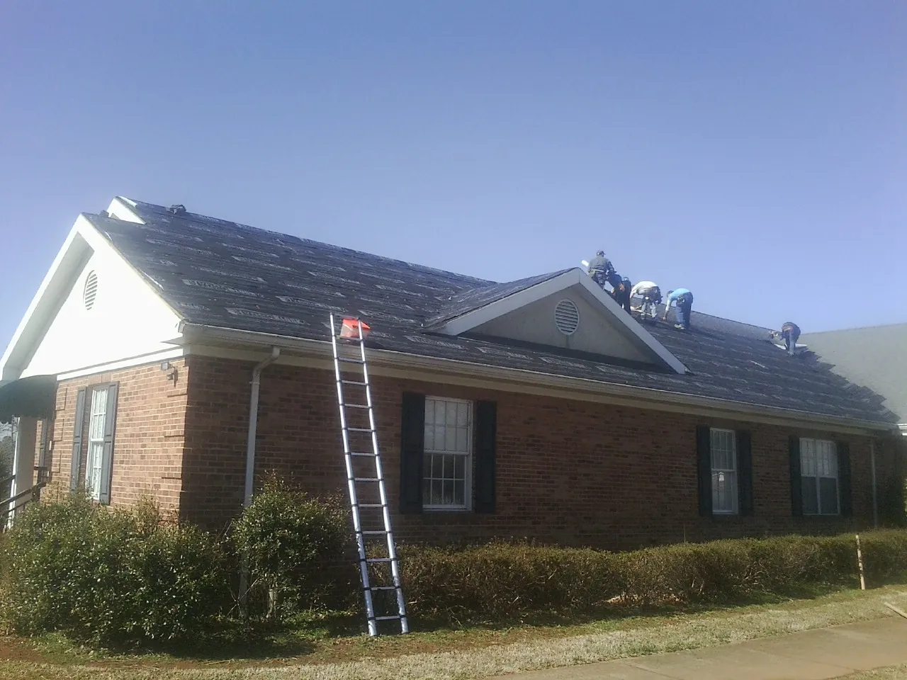 Crew tearing shingles off a brick ranch roof with a ladder set — Emergency Roof Repair work by our crew in Carney