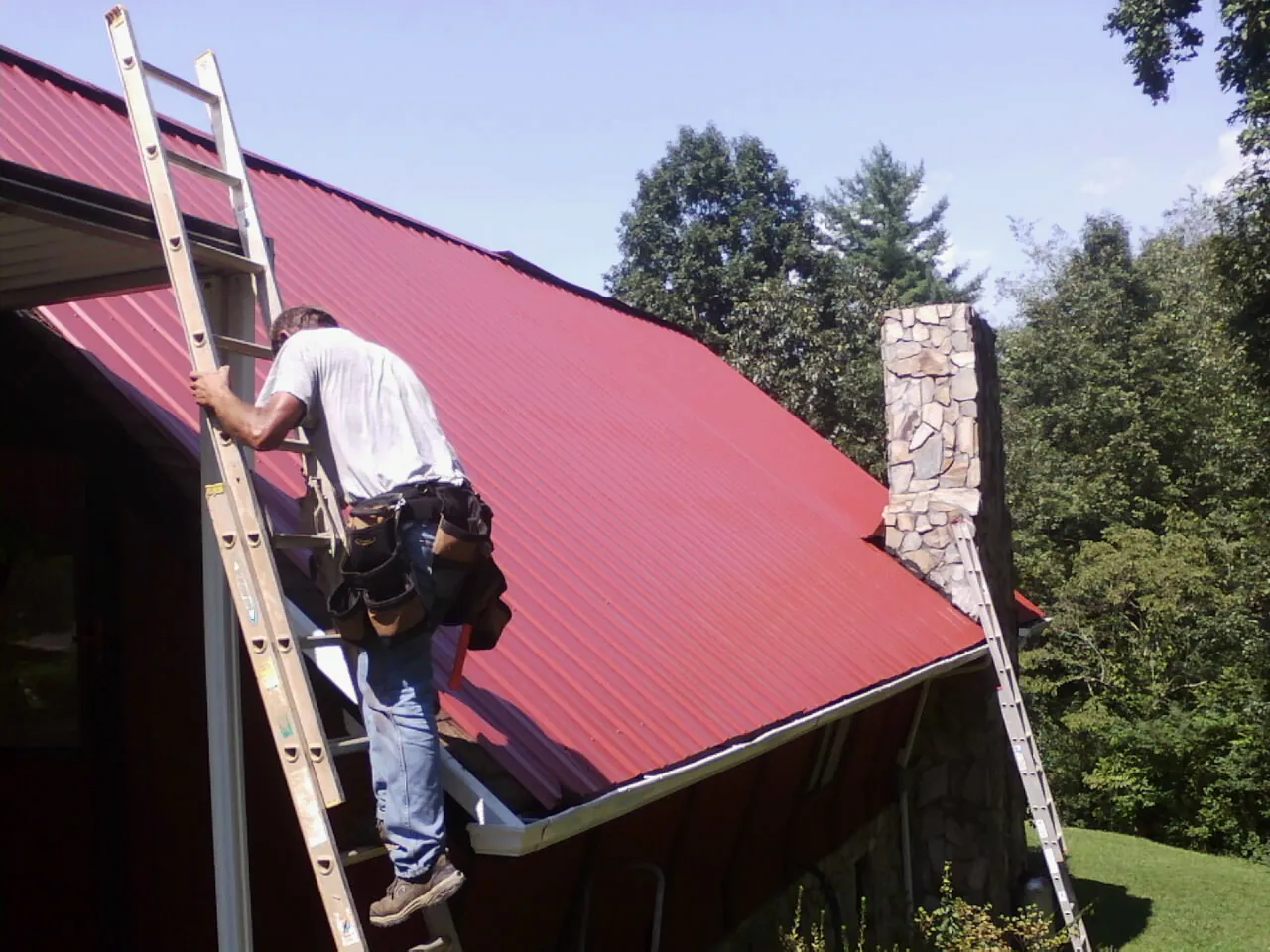 Roofer on a ladder beside a red metal roof and stone chimney — Soffit & Fascia Repair by Roofing Brothers in Carney